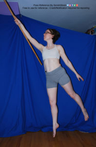 White woman holding a staff, pointing up at the upper left hand corner and jumping/floating.