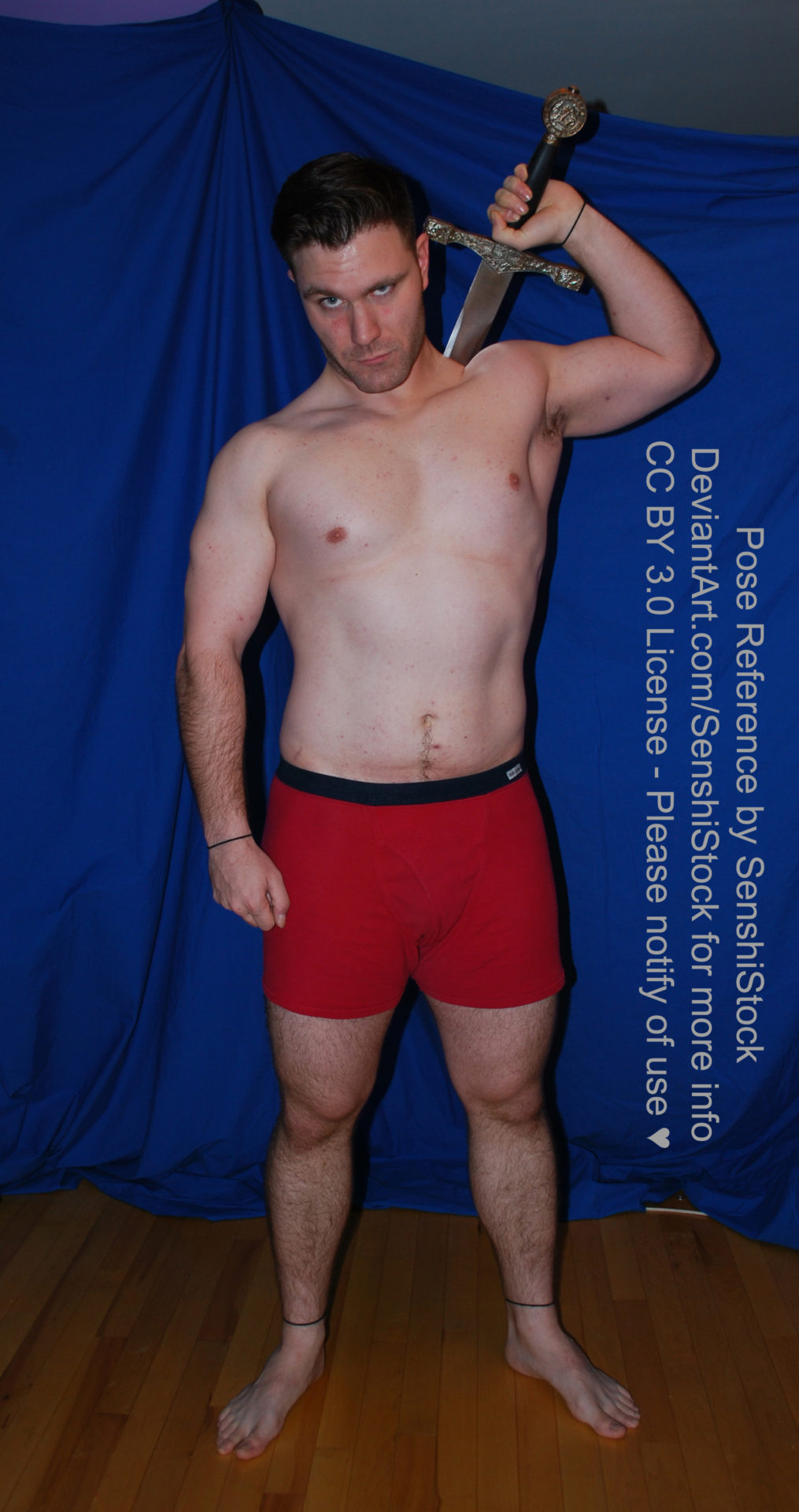 A muscular white man wearing a pair of red boxers is standing mostly straight up and relaxed, with one arm raised and holding a sword just barely shown from behind his back.