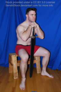 A muscular white man wearing a pair of red boxers is sitting on a wooden stool with his legs spread apart, arms raised and resting on the hilt of a sword. His chin is on his hands as he looks off into the distance.