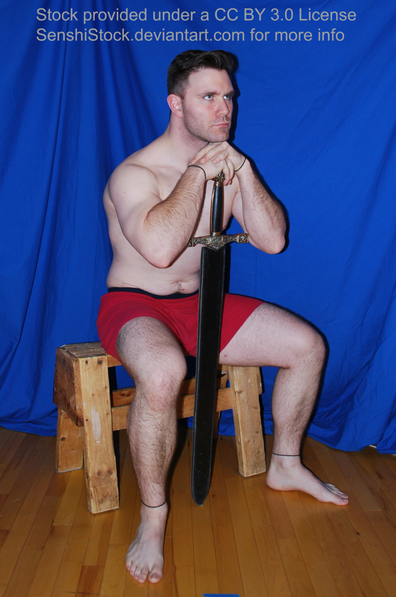 A muscular white man wearing a pair of red boxers is sitting on a wooden stool with his legs spread apart, arms raised and resting on the hilt of a sword. His chin is on his hands as he looks off into the distance.