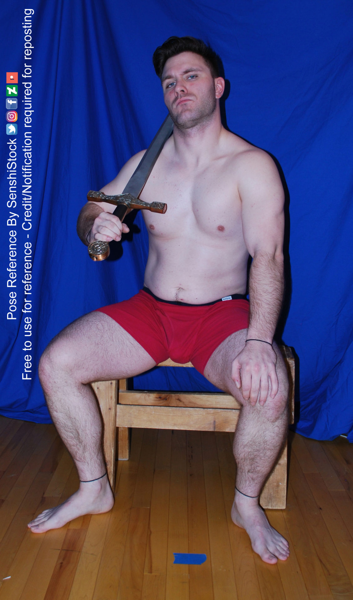 A muscular white man wearing a pair of red boxers is sitting on a wooden stool with his legs spread apart, one arm on his knee, the other holding up a sword that's resting on his shoulder. He's lifted up his head as he looks at you, as if challenging you.