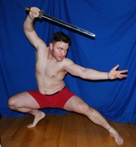 A muscular white man wearing a pair of red boxers is squatting, one leg straight out, the other bent at the knee and supporting his weight. One arm is out-stretched with palm up and fingers splayed, the other is holding up a sword above his head.