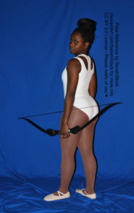 Thin black woman wearing a white one piece skin-tight bodysuit is standing, body facing away, while wearing pointe shoes, is holding a bow down at her side. She's looking back at the camera.