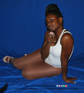Thin black woman wearing a white one piece skin-tight bodysuit is relaxed on the ground, legs back behind her, one arm holding her up, as the other is holding a gun pointed directly at the camera. She's looking at you with a soft but confident smile.