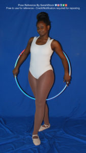 Thin black woman wearing a white one piece skin-tight bodysuit and pointe shoes is standing facing the camera. She's holding a hula hoop behind her, arms down, with one leg on either side.