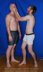 Two white men with boxers standing, one with both hands clasped on the cheeks of the other and squishing his face.
