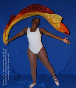 Thin black woman wearing a white one piece skin-tight bodysuit and pointe shoes is standing with her body facing the camera. One leg is straight down and holding her weight, the other is out on its toes. She is flinging fabric around her head, making a semi-circle, and looking up, arms spread apart.