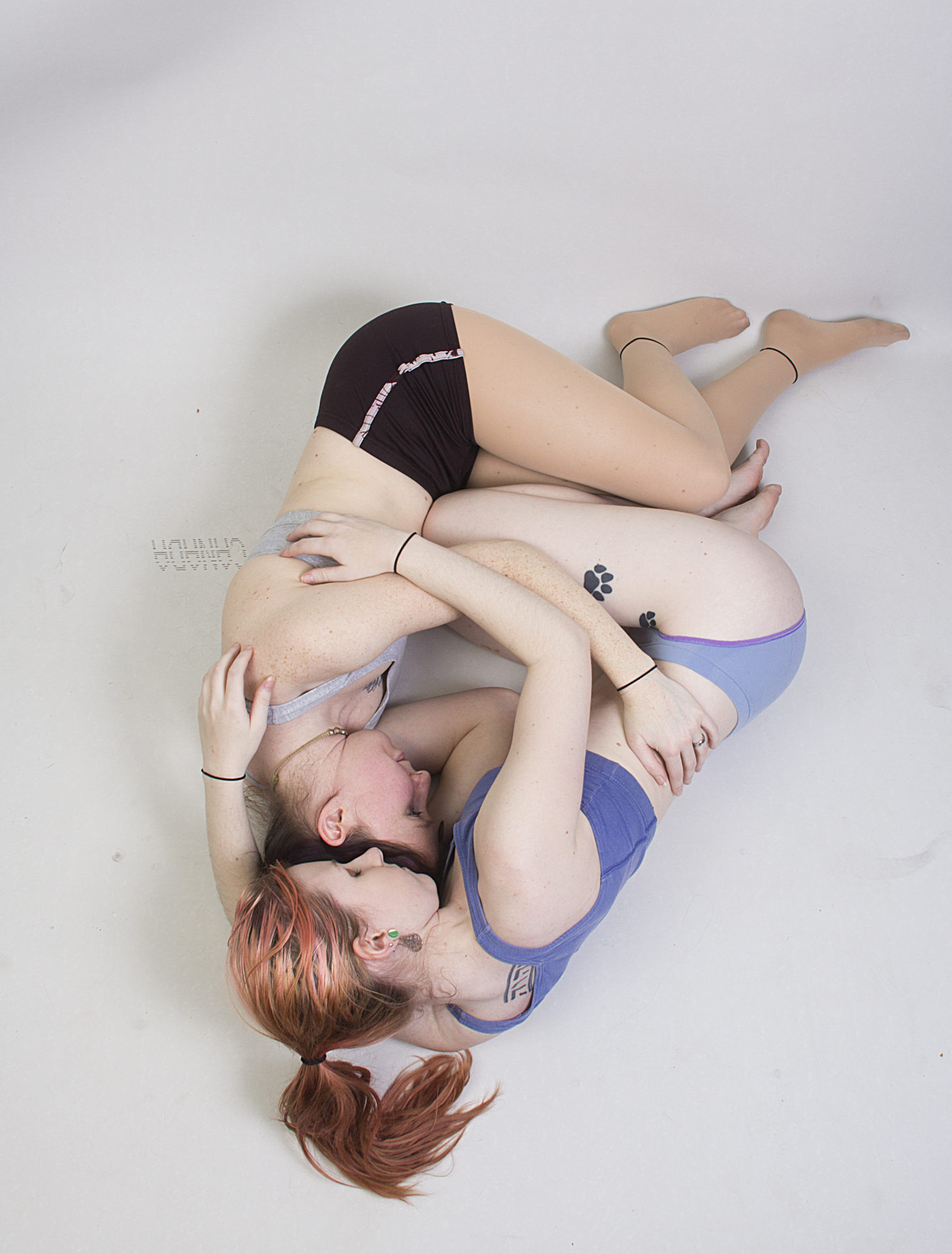 Two thin, white, female models wearing sports bras and short shorts, seen from above as they're laying on the floor in an embrace, legs bent and up against each other, and arms wrapped around each other. Their eyes are closed and they look relaxed. The shot has their feet at the top and heads at the bottom, both looking directly at the camera.