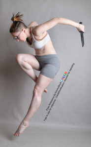 Thin white woman wearing white sports bra and form-fitting grey shorts and glasses, caught in mid-air leap downward as she holds a blade in each hand, arms pulled backward (only left arm can be seen). One leg is extended, the other is bent upward toward the chest.