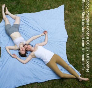 Two thin white women seen from a bird's eye view laying down with their heads together and holding hands.