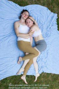 Two thin white women seen from a bird's eye view laying down cuddling with each other, legs and arms intertwined.
