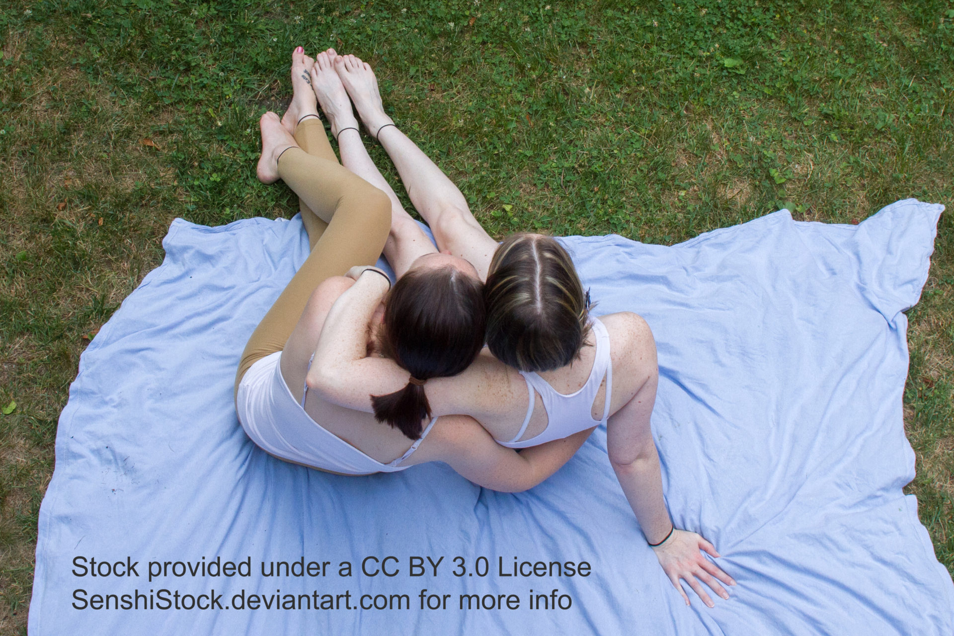 Two thin white women seen from a bird's eye view with them sitting on the ground, backs facing the camera, and cuddled up to each other.