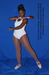Thin black woman wearing a white one piece skin-tight bodysuit and pointe shoes is standing facing the camera, legs apart, arms crossed in front of her, one in front of her stomach, the other by her neck and holding a magical crystal wand.