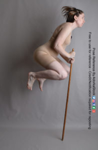 Thin white woman facing to the right jumping, her legs are bent in the air and the bottom of the stick is touching the ground.