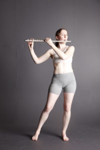White woman on a grey background playing a flute.