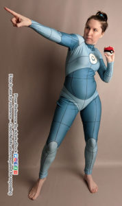A white woman wearing a teal suit standing and holding a pokeball, pointing up and off to the upper left.