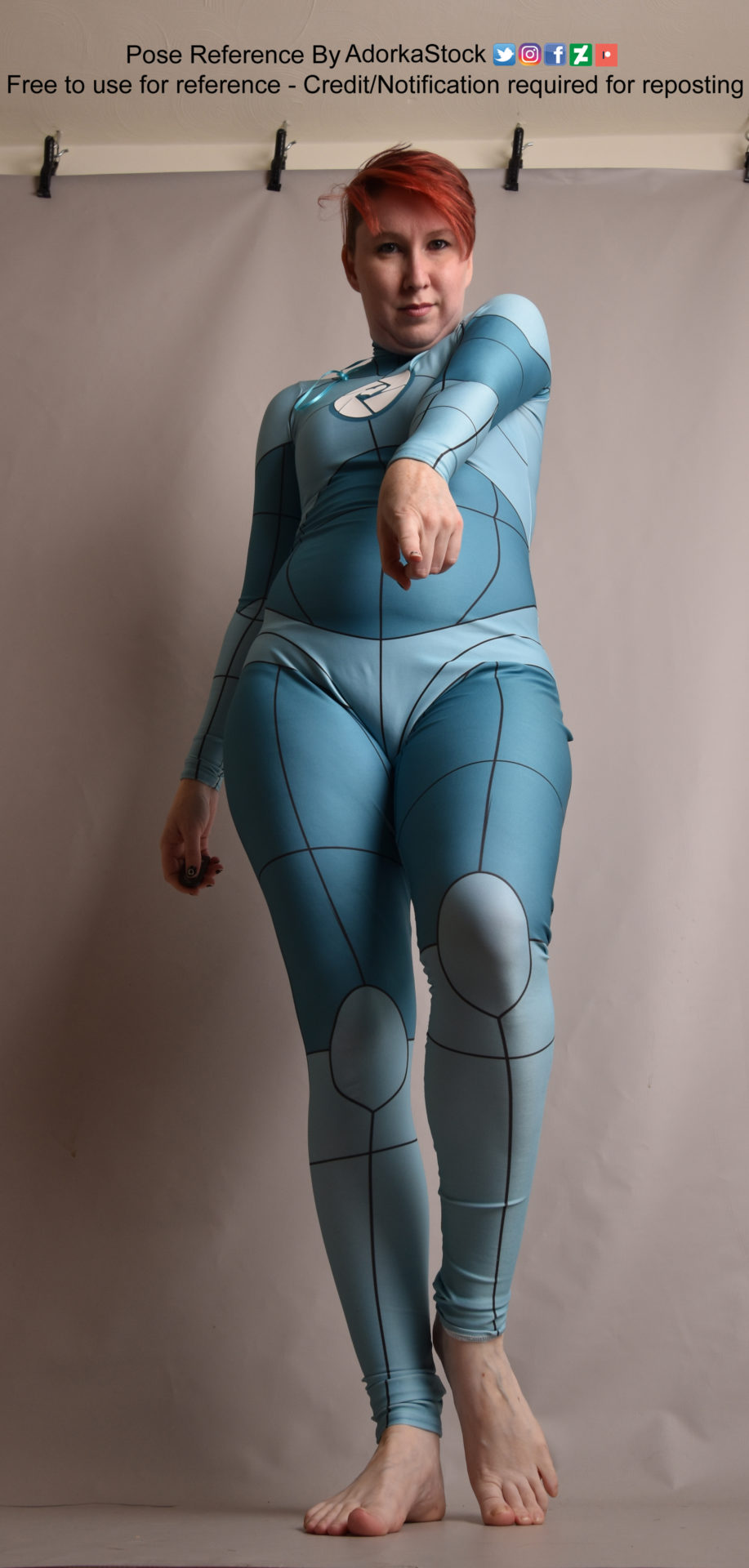 A person wearing an aqua colored body suit with grid lines on it, taken from a low angle, pointing up. The person is facing the camera and pointing their left finger accusingly at the camera.
