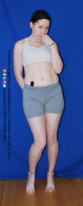 Thin, white, female model in coy, playful pose reference, one hand behind her back, one up by her face.