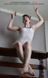 White woman sitting down, legs crossed, arms up in the air and doing the "devil horns" gesture with both hands, yelling.