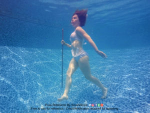 A underwater shot of a thin white woman in a bathing suit, holding a staff, acting like she's walking.