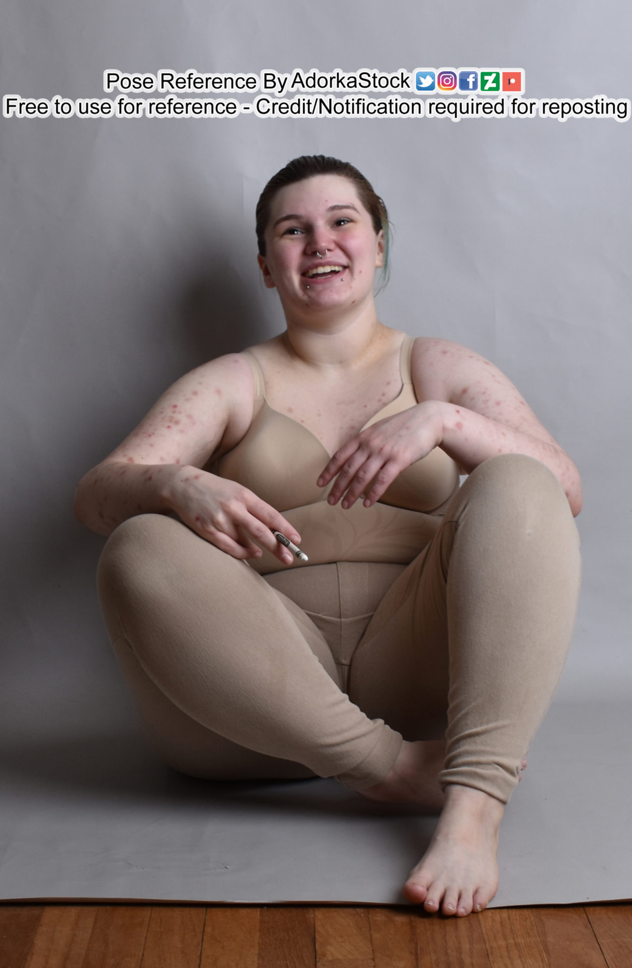 Pose reference model sitting on the ground with feet closer to the camera, arms resting on the knees, one hand holding a crayon like a cigarette. The model is leaning back and laughing a bit.