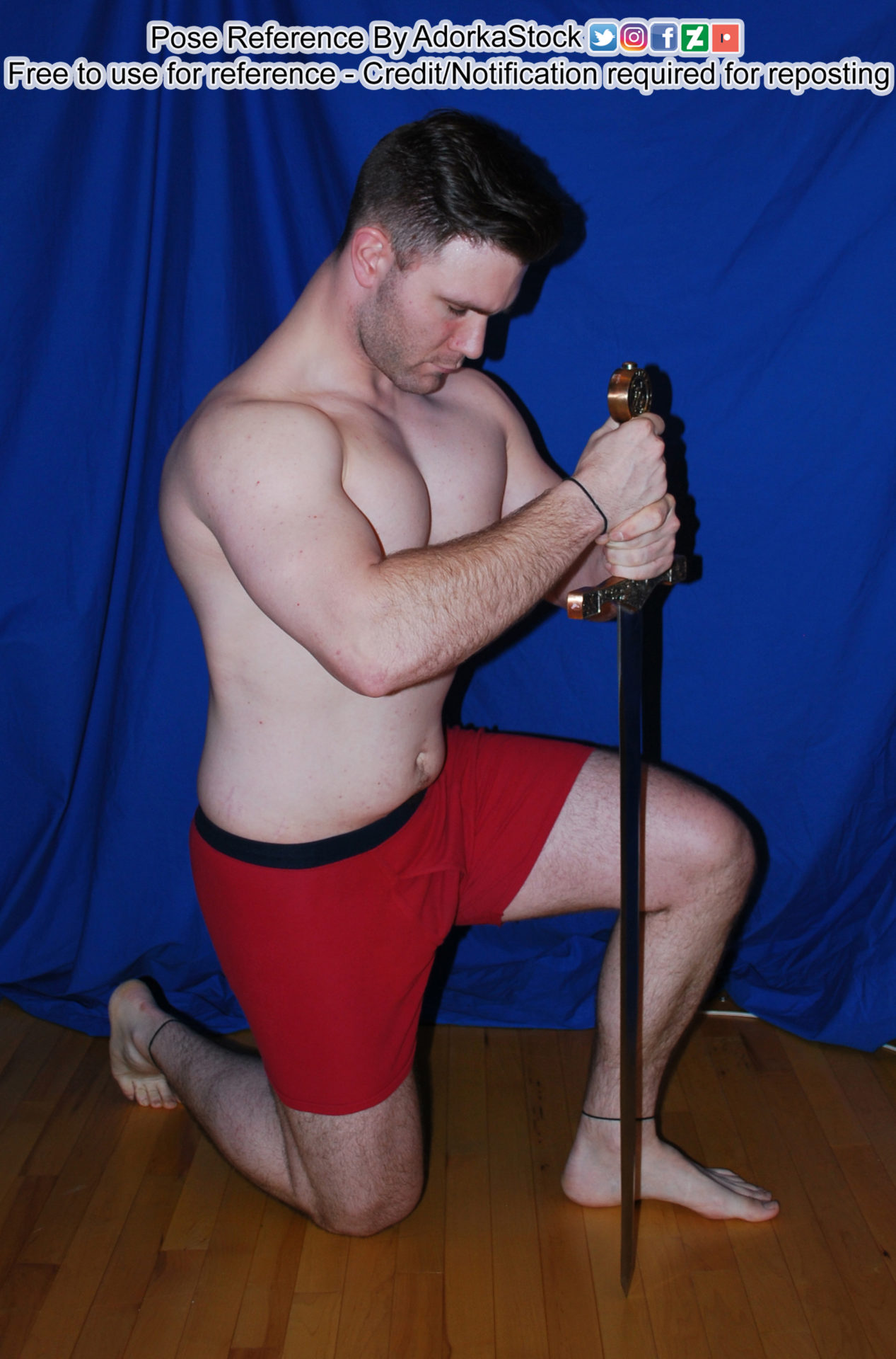 Muscular, white, male model on one knee holding the hilt of a sword with the tip placed on the ground. His head is bowed.