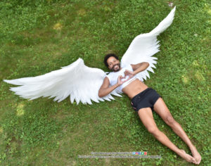 Thin, Latino, male model lying on the ground with huge white wings expanding out behind him, legs loose, hands shaped like a heart near his chest. The photo is taken from a high perspective.