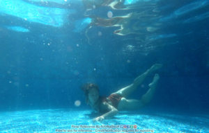 A underwater shot of a thin, fit woman, swimming with her chest against the bottom of the pool, legs up behind her and floating, with one arm reaching out on the pool floor.