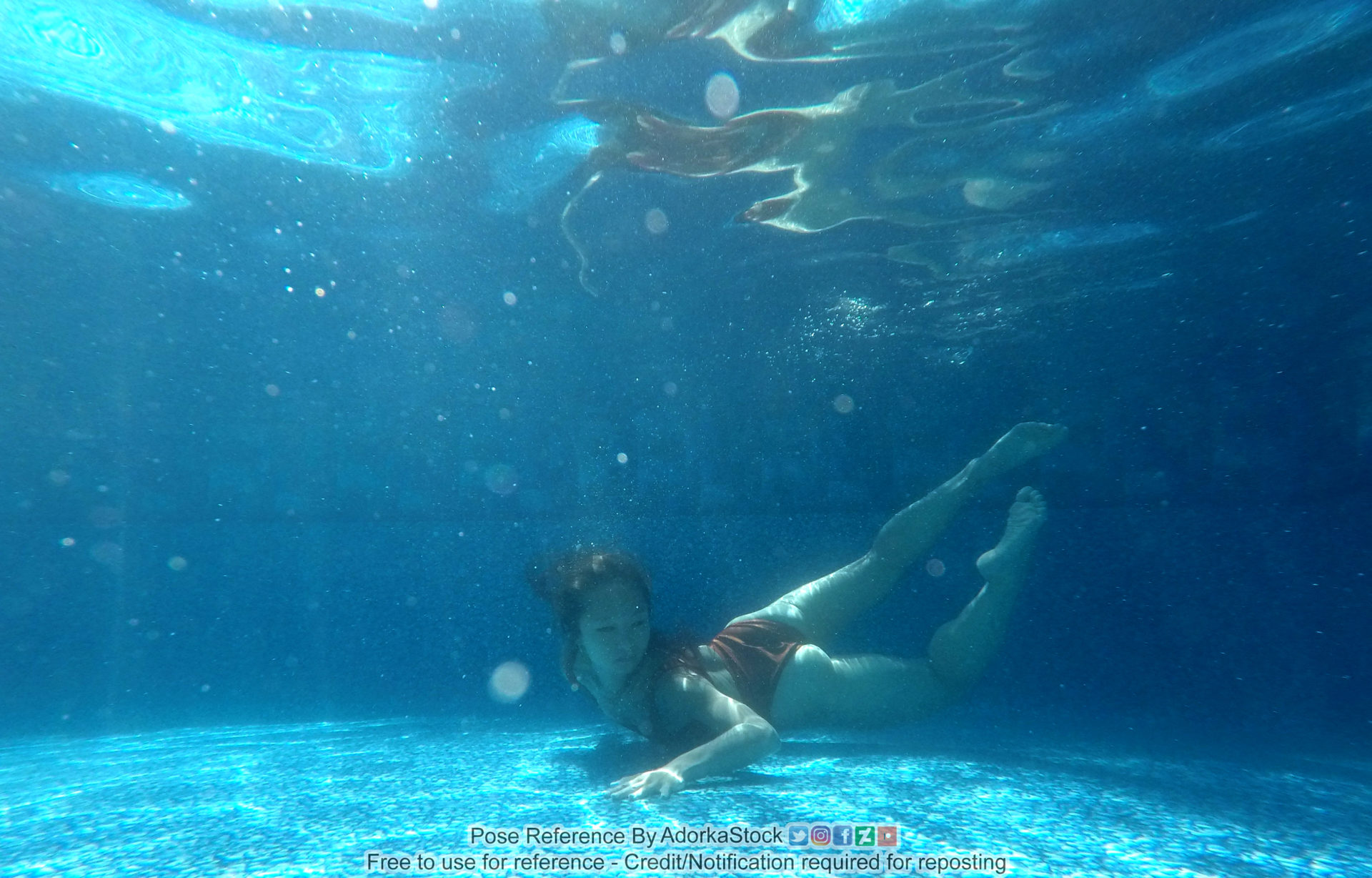 A underwater shot of a thin, fit woman, swimming with her chest against the bottom of the pool, legs up behind her and floating, with one arm reaching out on the pool floor.