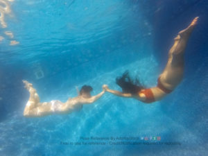 Two thin women are floating underwater, seen in profile, with their legs relaxed behind them, arms out stretched and grasping each others' hands.