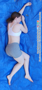 Thin, white, female pose reference model lying down from a high overhead perspective with her back mostly towards the camera