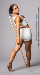 Fit, Latina, female model stepping away from the camera but leaning back with her head over a shoulder, arm swinging behind her holding a staff