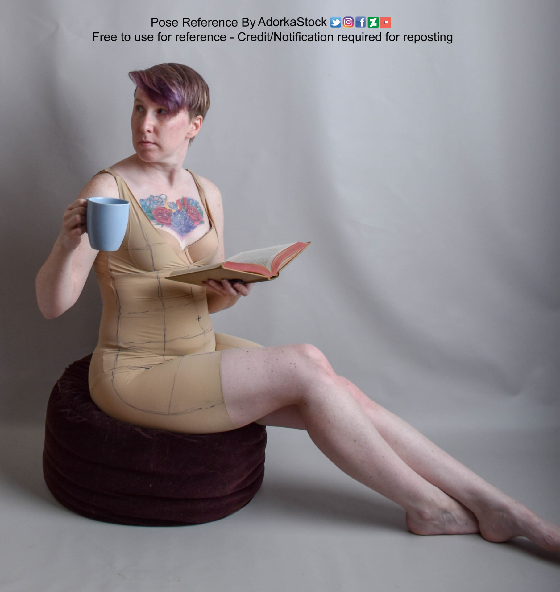 Thin, white, female pose reference model sitting on a cushion with legs to the side, holding a book and a mug