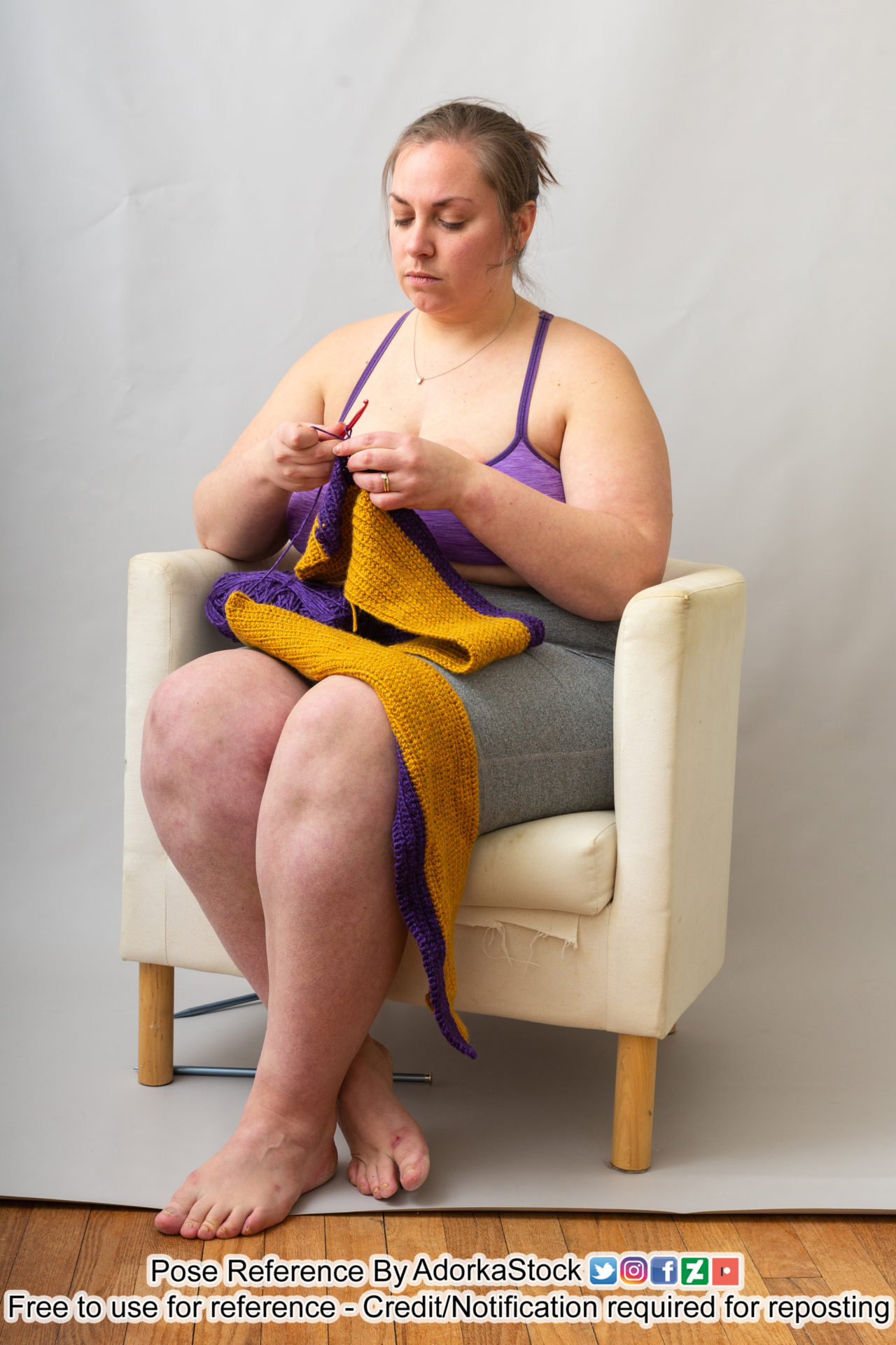 A woman in a purple sports bna and grey shorts sitting in a low backed beige arm chair crocheting a purple and yellow scarf. Her feet are crossed and she looks relaxed