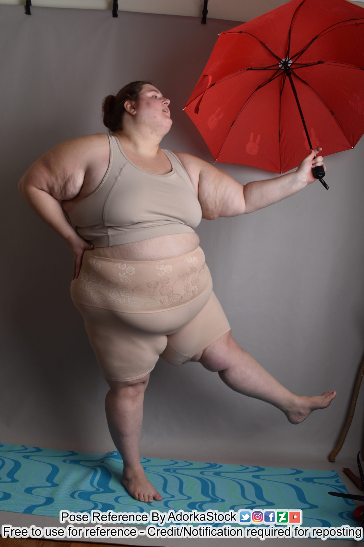 A heavyset woman in a grey sports bra and nude colored shapewear shorts and holding a red umbrella with white rabbits on it off to the side in her outstretched left hand. Her left leg is raised and out to the side. Her right hand is on her hip and her mouth is open like she's singing and dancing