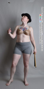 A woman in a tan bra and grey shorts wearing a black fedora, holding a brown stick in her left hand and flipping a large coin with her right. The coin is in the air