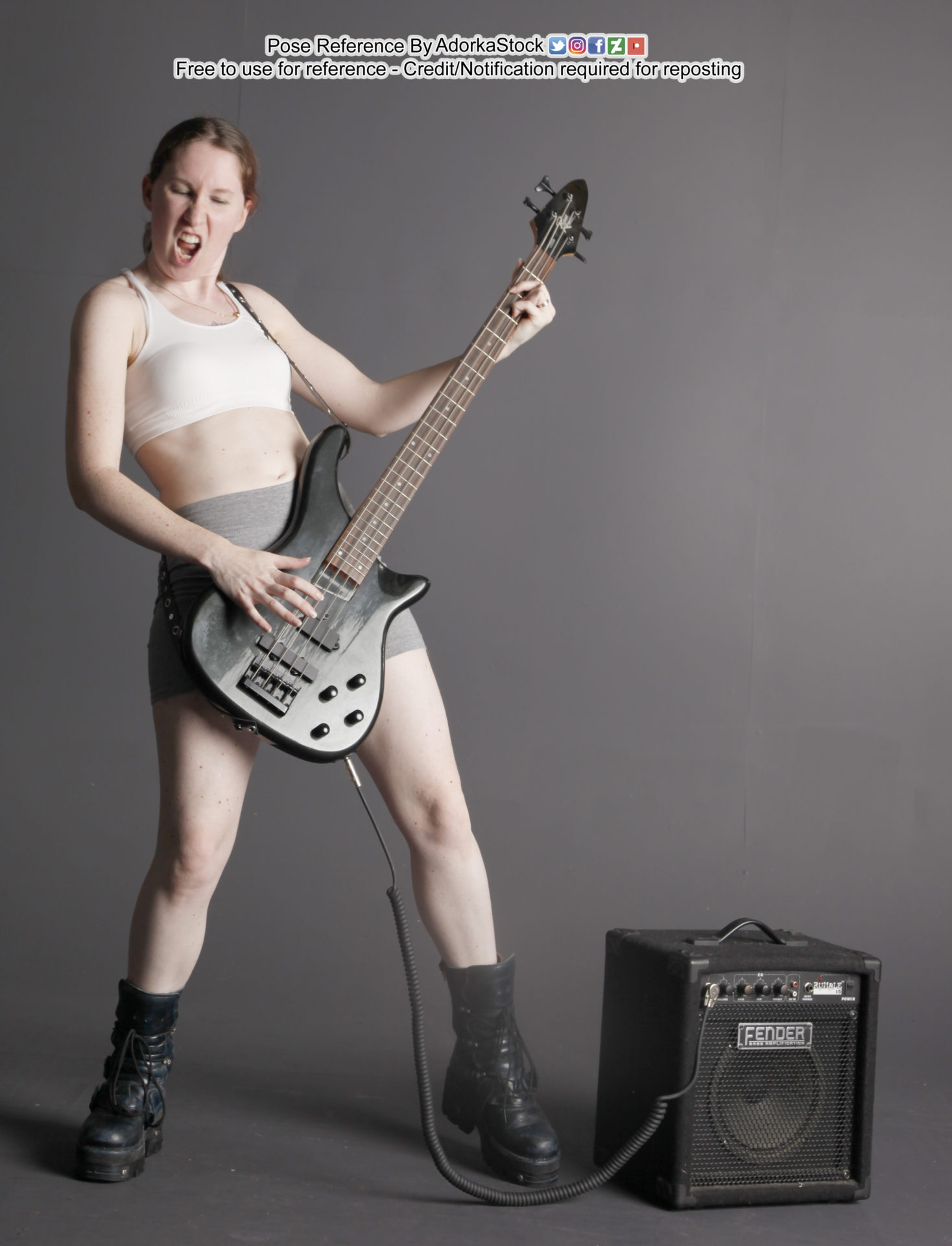 A woman in a white sports bra, grey shorts and black combat boots playing a black Fender guitar hooked up to a black Fender amp with a curly cord. Her mouth is open and her eyes closed as if in mid song