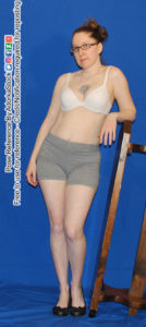 A woman in a white bra, grey shorts and black wedge heels leaning casually with her left arm propped up on an upturned dark brown wooden bench with a smirk on her face. Her right arm is by her side