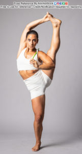 A woman in a white sports bra and shorts facing the camera, left leg stretched up above her head being held with her right hand