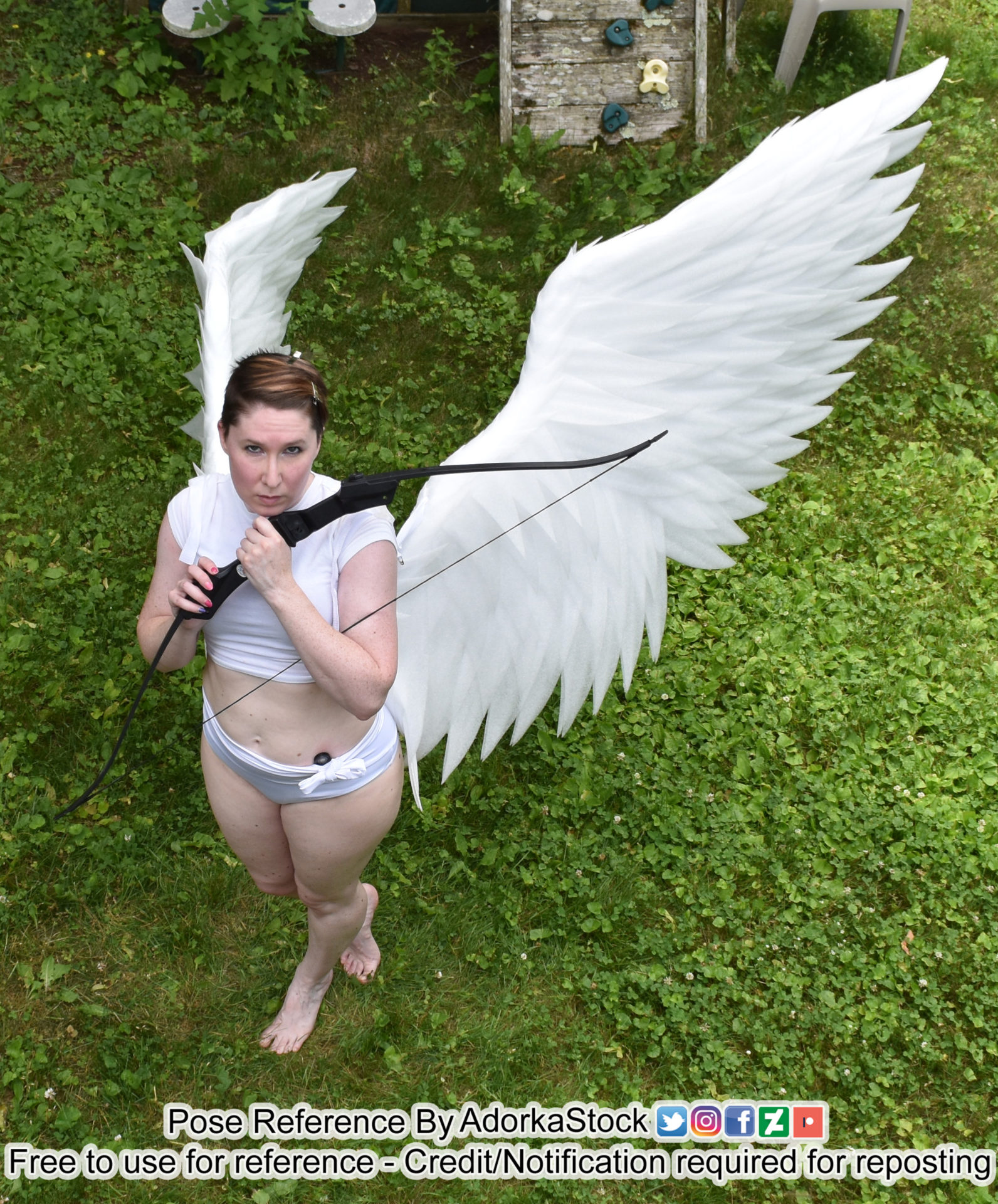 A picture taken from a high angle of a woman in a white swim suit with a crop top, wearing large white angel wings and clutching a black bow weapon close to her chest and looking up