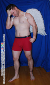 A muscular man wearing red boxer briefs and a pair of small angel wings with his head angled down and eyes closed with his forehead in his raised right palm and his fingers in his hair. His left hand is clenched into a fist