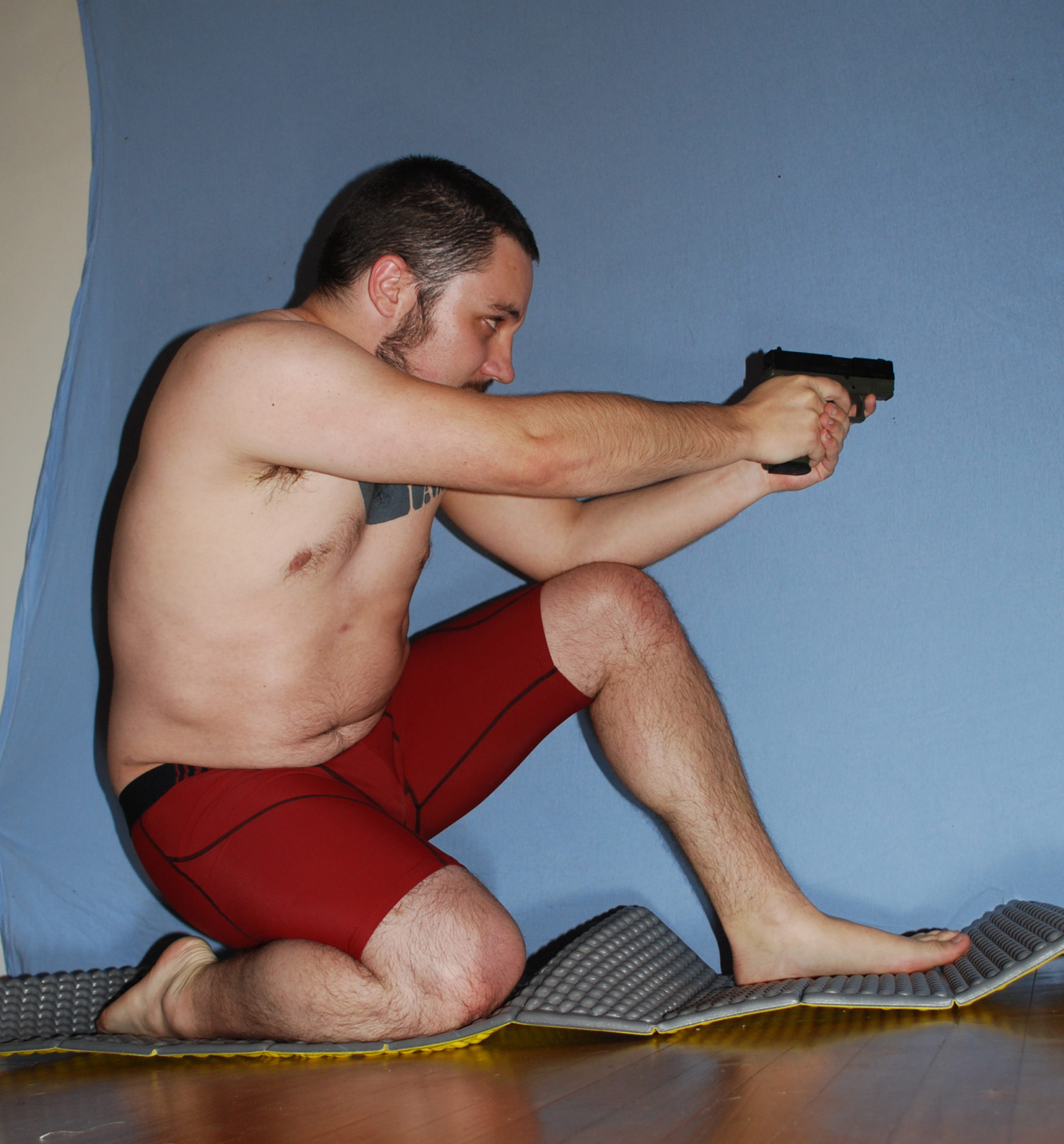 A man in red boxer briefs kneeling on his right knee. His left leg is out in front and bent with the knee holding up his left elbow. He's holding a pistol in both outstretched hands and pointing at a target off camera to the right