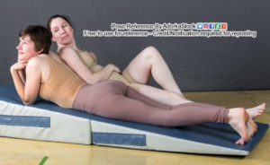Two women lying on a padded exercise incline that has the highest end facing to the left. The woman in the front is wearing a tan shirt and brown leggings, is smiling and lying on her belly with her chin propped up on her hands. The woman behind her is wearing a nude colored bodysuit and has her elbow on the other woman's back. She is smiling at the back of the other woman's head