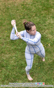 A high angle picture of a woman in a white line suit facing the right with her left leg behind her and her arms raised above and behind her head, brandishing a katana with a white handle and hilt
