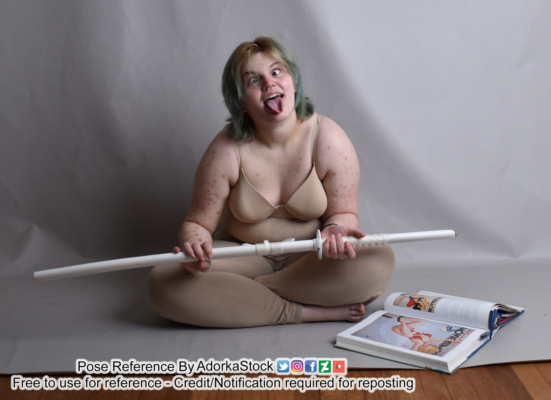 A person in a nude colored top and skinny jeans sitting crosslegged with a white handled katana across their knees and in their hands. The person has their eyes crossed and tongue sticking out. There is a blue book on the right with pictures of pinup girls in it