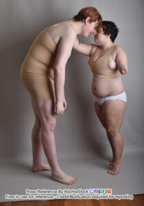 Very tall female model leaning down with arms limp to press her forehead against a very short male model who is on tip toes, reaching up with his far arm. The near arm is partially amputated near the elbow.