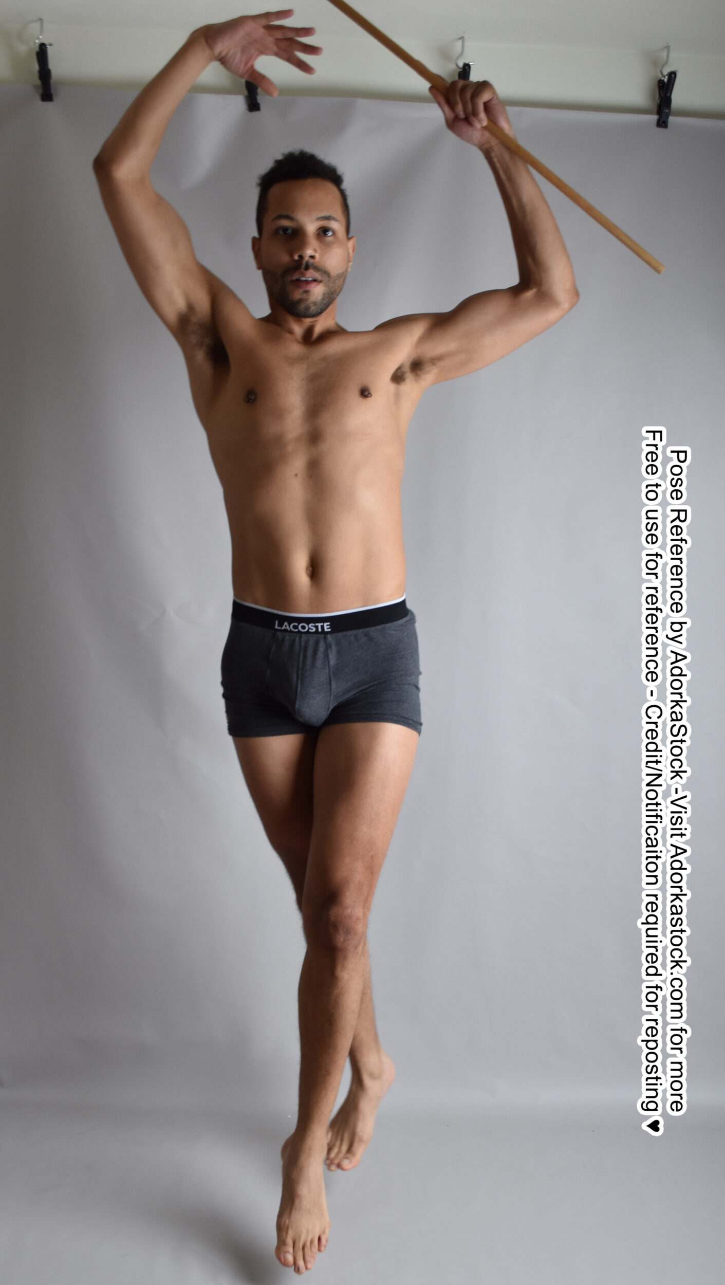 Thin, Latino, male pose reference model in jumping pose with toes pointed, hands over head, holding a staff.