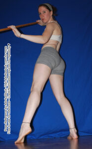 Thin, white, female pose reference model in low angle, back facing standing pose with staff held up chest height to the side.