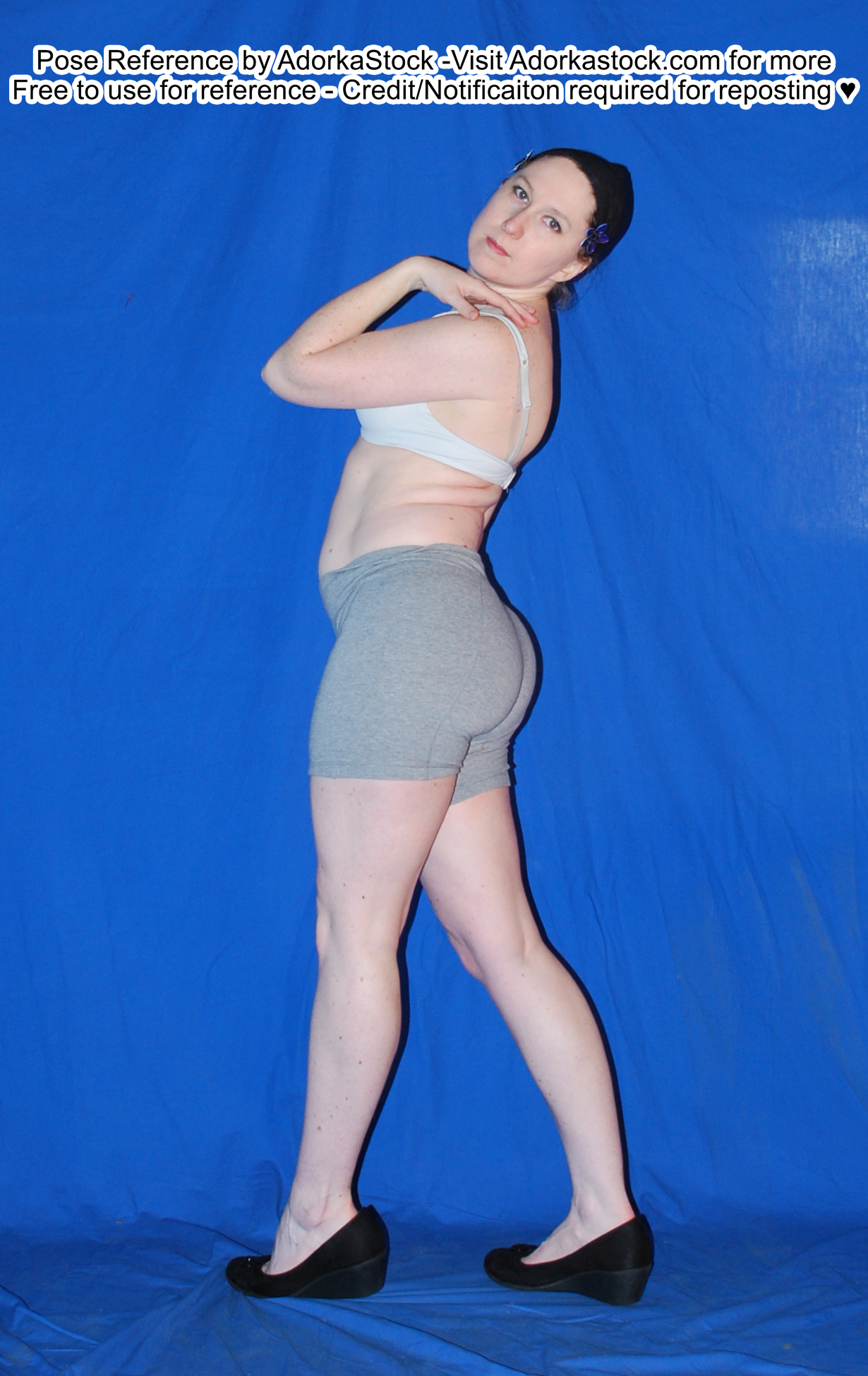 Thin, white, female pose reference model in standing profile pose with one hand up by her face, looking over her shoulder at the camera.
