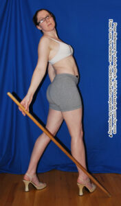Thin, white, female pose reference model in 3/4 back pose with full body twist, one hand on a staff resting on the ground.